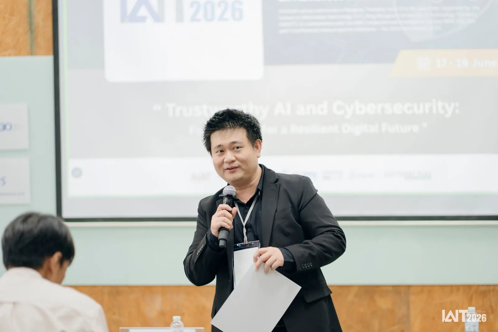Charnsak Srisawatsakul speaks into a microphone in front of the IAIT 2026 conference screen